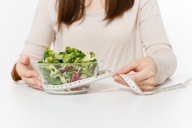 Nahaufnahme abgeschnittene Frau am Tisch mit grünem Salat in Glasschüssel, Maßband des Schneiders isoliert auf weißem Hintergrund. Richtige Ernährung, vegetarisches Essen, gesunder Lebensstil, Diätkonzept. Platz kopieren.