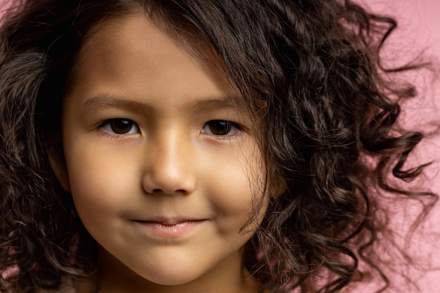 Nahaufnahme abgeschnittener Kopfschuss des reizenden Kindes, kleines Mädchen mit dunklem lockigem Haar, brauner Haut, braunen Augen, sanftem Lächeln, direkt schauend. Kinder, Kinder, Kindheitskonzept.