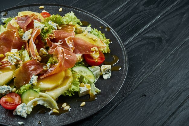 Nahaufnahme Ansicht auf Salat mit Honigbirne, dor Blauschimmelkäse und Schinken auf stilvollem dunklem Teller auf schwarzem Holztisch. Selektiver Fokus