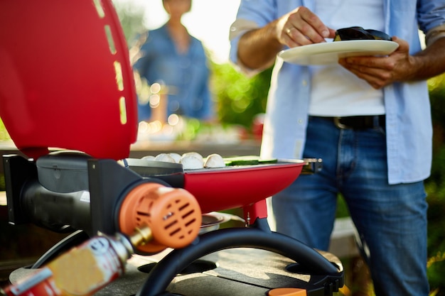 Nahaufnahme auf der Hand des Mannes, die Gemüse auf dem Grillgasgrill im Freien im Hinterhof röstet, Gemüse auf dem Grill, Sommerfamilienpicknick, Essen in der Natur.