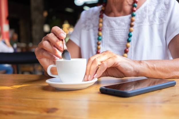 Nahaufnahme auf der Hand einer älteren Frau, die eine weiße Kaffeetasse hält, während sie sich für eine Pause im Coffeeshop, Holztisch und Mobiltelefon entspannt