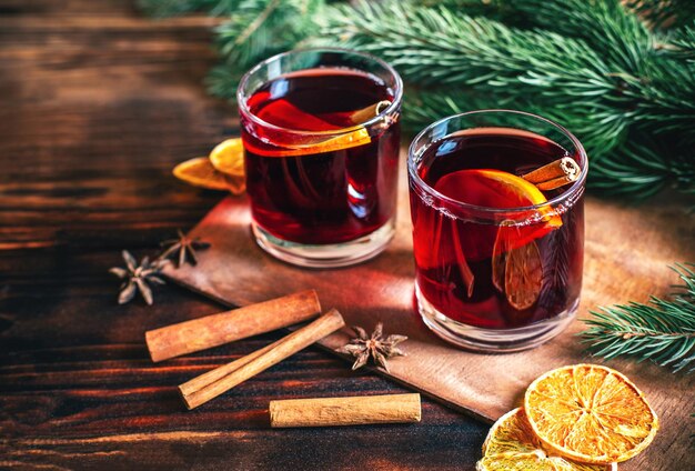 Nahaufnahme auf einem heißen alkoholischen Getränk Glühwein in zwei transparenten Gläsern mit Zimtstange, Orange und Sternanis auf einem dunklen Holztisch vor dem Hintergrund von Weihnachtsbaumzweigen