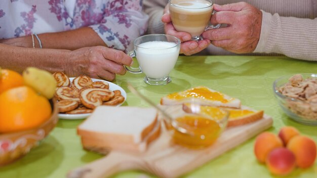 Nahaufnahme auf erwachsenen Seniorhänden, die eine Milch- und Cappuccinotasse halten, während sie zu Hause am Tisch frühstücken