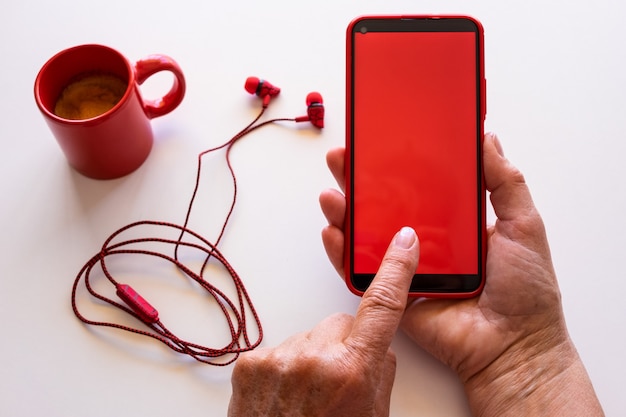 Nahaufnahme auf weiblichen Händen mit Telefon mit rotem Bildschirm. Weißer Desktop mit Kaffeetasse und Kopfhörern