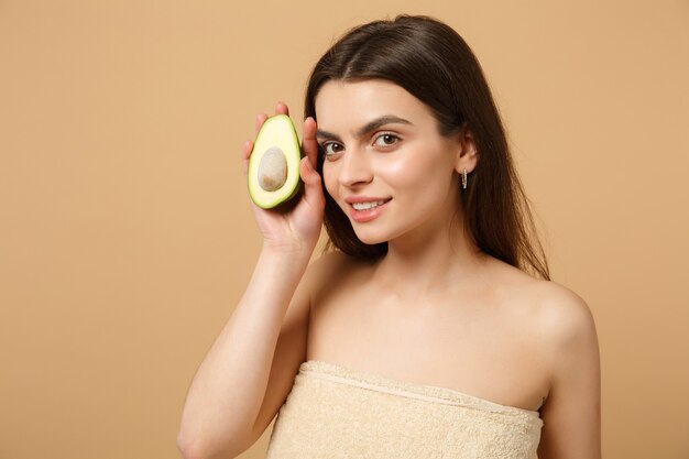 Nahaufnahme brünette halbnackte frau mit perfekter haut nackt make-up hält avocado isoliert auf beige pastellfarbener wand