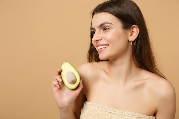 Nahaufnahme brünette halbnackte Frau mit perfekter Haut nackt Make-up hält Avocado isoliert auf beige pastellfarbener Wand