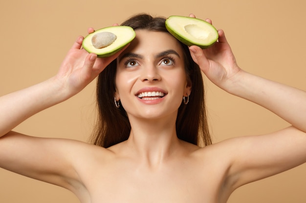 Nahaufnahme brünette halbnackte Frau mit perfekter Haut nackt Make-up hält Avocado isoliert auf beige pastellfarbener Wand