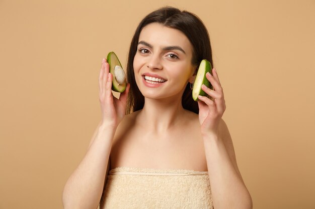 Nahaufnahme brünette halbnackte Frau mit perfekter Haut nackt Make-up hält Avocado isoliert auf beige pastellfarbener Wand