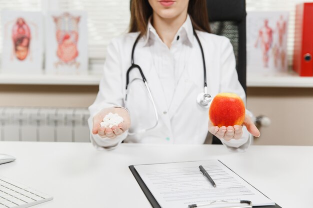 Nahaufnahme der abgeschnittenen Ärztin, die am Schreibtisch sitzt und eine Flasche mit weißen Pillen in der Handfläche hält, roter Apfel im hellen Büro im Krankenhaus