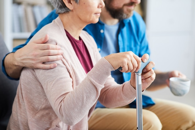 Nahaufnahme der älteren Frau, die Krücke hält und auf dem Sofa mit männlicher Krankenschwester sitzt, die sie umarmt und in der Nähe sitzt