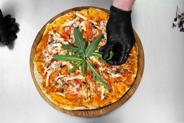 Nahaufnahme der behandschuhten Hände des Küchenchefs, die Pizza mit Grüns, Rucola dekorieren.