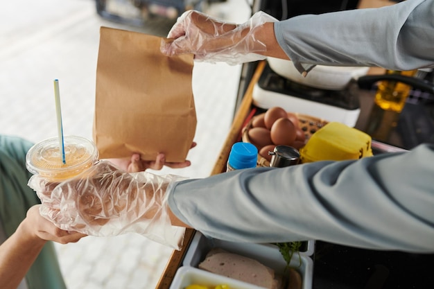 Nahaufnahme der behandschuhten Hände eines LKW-Verkäufers, der Limonade und verpacktes Fast Food vorbeireicht