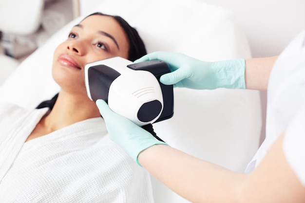 Nahaufnahme der Frau, die sich einer Hautanalyse mit Hilfe eines modernen Werkzeugs in der Nähe ihres Gesichts unterzieht