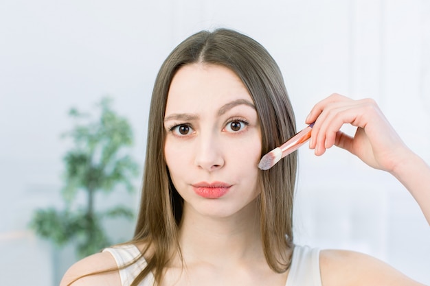 Nahaufnahme der glücklichen jungen Frau Gesicht mit Make-up Pinsel auf hellem Hintergrund. Schönheitsmädchen-Hautpflegekonzept. Speicherplatz kopieren