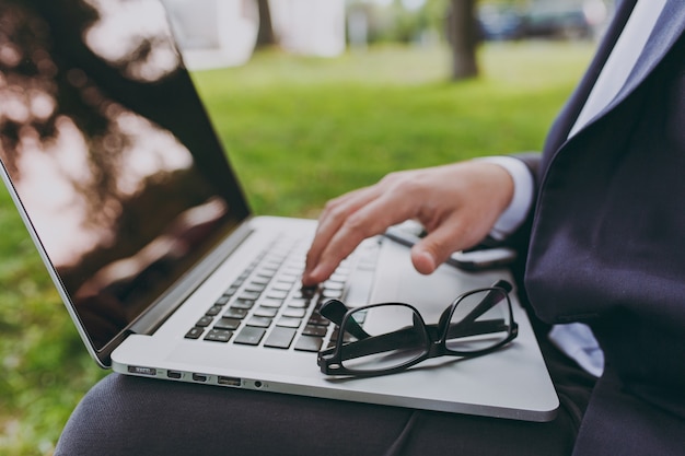 Nahaufnahme der Hände auf der Tastatur abgeschnitten. Geschäftsmann im klassischen Anzug, Brille. Mann sitzt auf weichem Hocker, arbeitet an Laptop-PC im Stadtpark auf grünem Rasen im Freien. Mobile Office-Konzept. Seitenansicht.