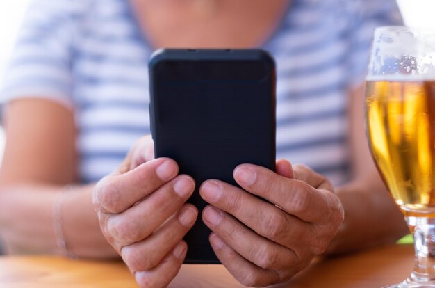 Nahaufnahme der Hände der Frau mit Telefon in der Nähe eines Glases eisgekühltes Bier Verschwommene kaukasische Dame, die mit alkoholischem Getränk am Restauranttisch sitzt