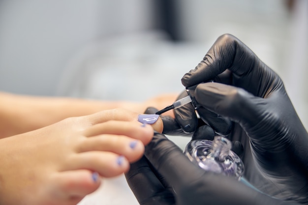 Nahaufnahme der Hände der Kosmetikerin in Latexhandschuhen, während sie die Zehennägel der Frau malt