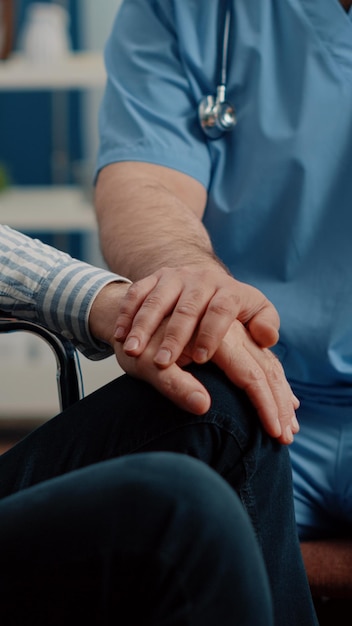 Nahaufnahme der Hände der Mannkrankenschwester, die älteren Patienten mit chronischer Behinderung im Pflegeheim tröstet. Medizinischer Assistent, der eine Person im Rollstuhl unterstützt. Spezialist hilft Erwachsenen