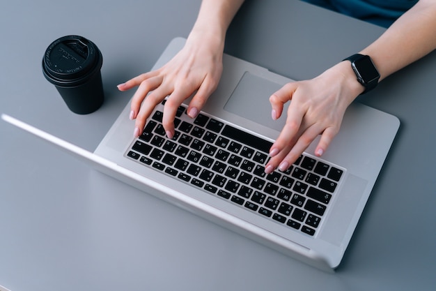 Nahaufnahme der Hände der nicht erkennbaren Geschäftsfrau, die auf der Laptoptastatur sitzt, die am Schreibtisch auf dem Hintergrund des Fensters im Büroraum, selektiver Fokus, Kaffeetasse sitzt.