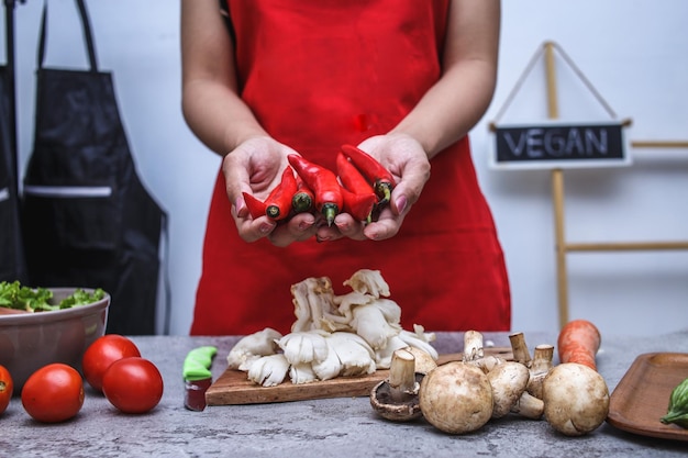 Nahaufnahme der Hände des Küchenchefs mit Schürze, die rotes Chili mit verschiedenen frischen Lebensmitteln auf dem Tisch hält.