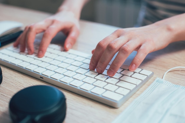 Nahaufnahme der Hände einer Frau, die auf einer modernen drahtlosen Tastatur tippt. Medizinische Maske an seiner Seite stockfoto