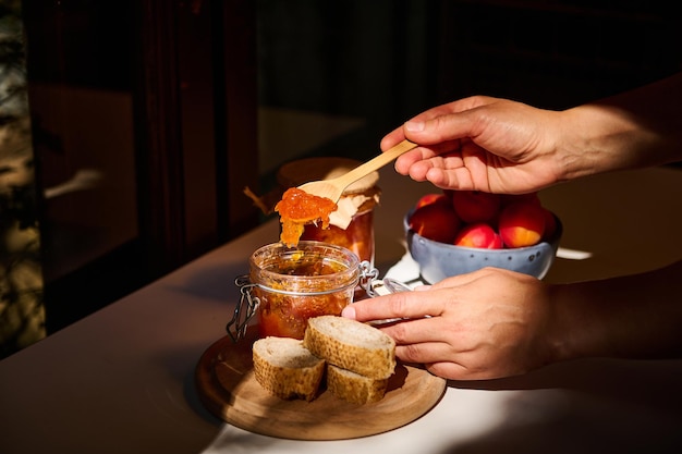 Nahaufnahme der Hände einer Hausfrau, die einen Holzlöffel hält und hausgemachte Aprikosenmarmelade auf Brot verteilt