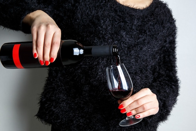 Nahaufnahme der Hände einer jungen Frau, die Rotwein in ein Glas aus einer Flasche gießt, rote Maniküre an den Fingern