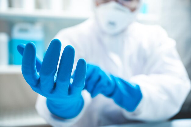 Nahaufnahme der Hände eines jungen, nicht erkennbaren Wissenschaftlers, Forschers und Arztes, der blaue Handschuhe mit weißem PSA-Kit und Uniform mit covid19-Gesichtsschutzmaske zur Sicherheit im Labor trägt