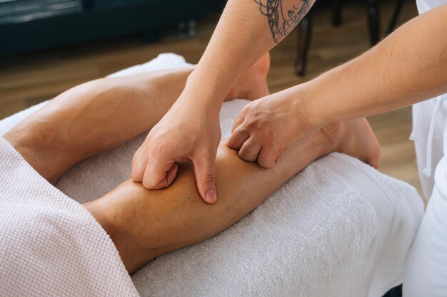 Nahaufnahme der Hände eines nicht erkennbaren männlichen Masseurs mit stark tätowierten Händen, die den Unterschenkel eines muskulösen Athleten nach dem körperlichen Sporttraining massieren und auf dem Bauch am Massagetisch liegen.