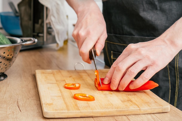 Nahaufnahme der Hände eines nicht erkennbaren Mannes, der rote Paprika mit einem Messer auf einem Holzschneidebrett in der Nähe eines Stahlsiebs hackt