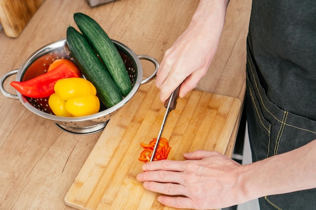 Nahaufnahme der Hände eines nicht erkennbaren Mannes, der rote Paprika mit einem Messer auf einem Holzschneidebrett in der Nähe eines Stahlsiebs hackt