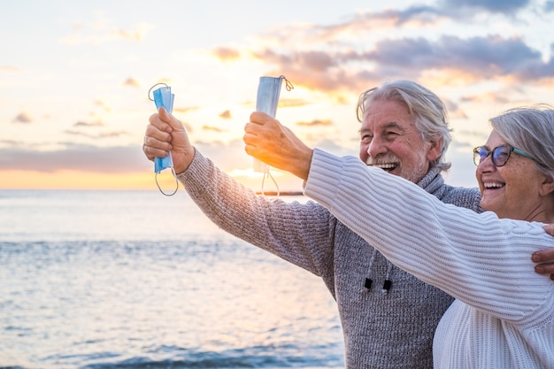 Nahaufnahme der Hände von zwei Personen, die sich mit ihrer Maske zum Schutz vor Covid oder Viren in den Händen halten, nachdem sie das Coronavirus gewonnen haben und am Strand frei im Freien sind