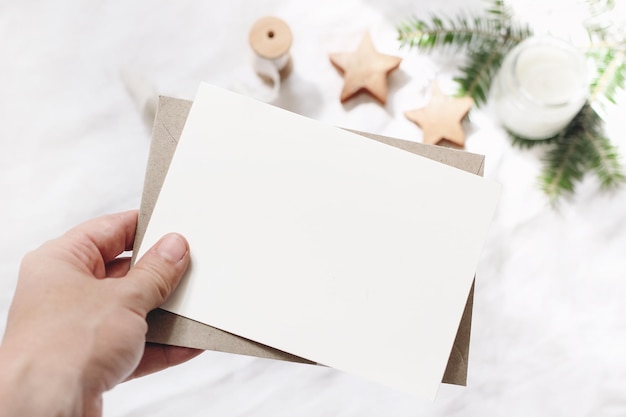 Nahaufnahme der Hand der Frau, die eine leere Papierkarte hält Grußkarten-Mockup-Szene Chritsmas Winterdesign Feminin gestaltet stock photo Der Hintergrund jedoch unscharf mit Tanne Zweig Kerze und Holzsterne