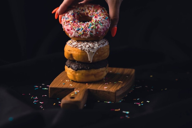 Nahaufnahme der Hand einer Frau, die einen köstlichen Donut von einem Holzbrett nimmt Köstliche Donuts, die auf einem hölzernen Schneidebrett auf schwarzem Hintergrund angeordnet sind