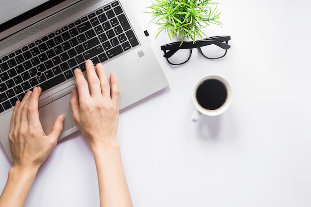 Nahaufnahme der Hand einer Person, die auf Laptop mit Kaffeetasse schreibt; Brillen und Blumentopf auf weißem Schreibtisch