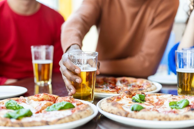 Nahaufnahme der Hand eines afrikanischen Kerls, der ein Glas Bier im Pizzarestaurant nimmt Multiethnische Freunde, die Spaß daran haben, Pizza zu essen und Bier im Pub zu trinken