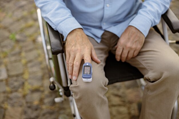 Nahaufnahme der Hand eines behinderten Mannes im Rollstuhl mit dem Fingerspitzen-Pulsoximeter zum Messen