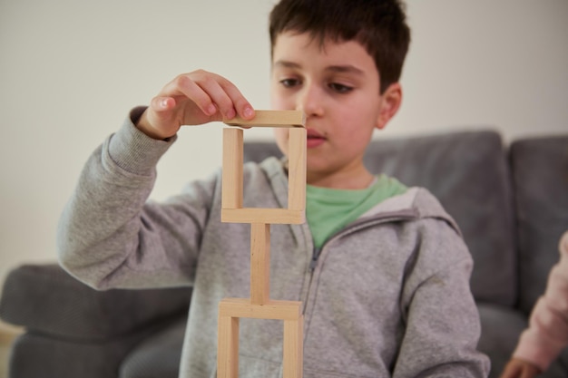 Nahaufnahme der Hand eines europäischen, gutaussehenden, verwirrten Schuljungen, der sich auf den Bau einer komplexen hohen Struktur aus Holzblöcken konzentriert. Feinmotorik, Konzentration und pädagogisches Freizeitkonzept