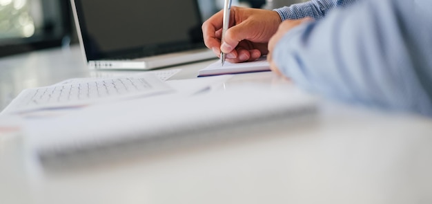 Foto nahaufnahme der hand eines geschäftsmannes, der notizen macht, indem er einen bleistift in einem modernen, hellen büro vor dem leeren dunklen bildschirm eines laptop-notebooks hält und das schreiben von notizen für ein vorstellungsgespräch plant