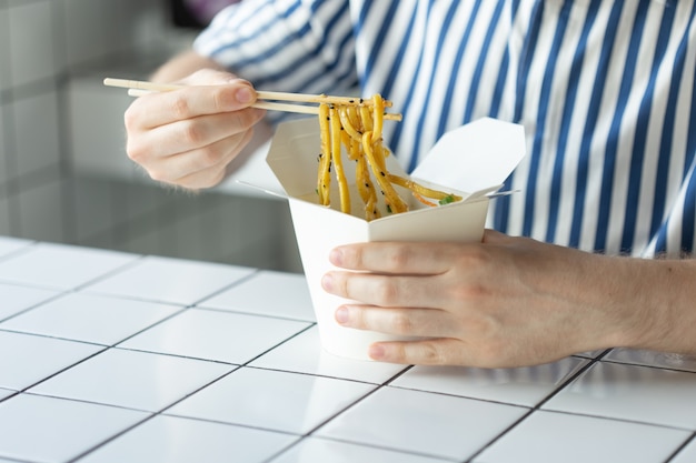 Nahaufnahme der Hand eines jungen nicht identifizierten Mannes in einem Hemd halten Holzstäbchen chinesische Nudel. Köstliches asiatisches Küchenkonzept.