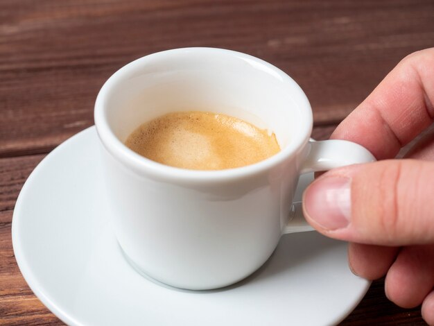 Nahaufnahme der Hand eines Mannes, der eine kleine weiße Tasse aromatischen Espressos über einer weißen Untertasse hält.