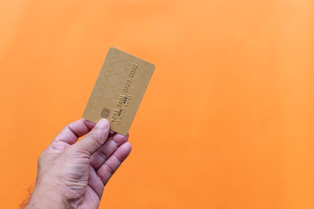 Nahaufnahme der Hand eines Mannes, der eine Kreditkarte auf orangefarbenem Hintergrund hält. Geschäftsmann zeigt Debitkarte, Online-Shopping, Zahlung, Online-Banking, Geldtransfer-Konzept, selektiver Fokus, Kopierraum auf der rechten Seite