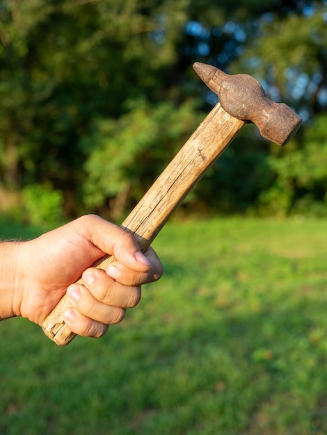 Nahaufnahme der Hand eines Mannes, die einen alten schmutzigen Hammer hält, unscharfer grüner Hintergrund Vertikales Foto