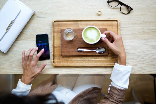 Nahaufnahme der jungen Frau, die Matcha-Grüntee-Latte auf Holztisch im Café trinkt. Ansicht von oben.