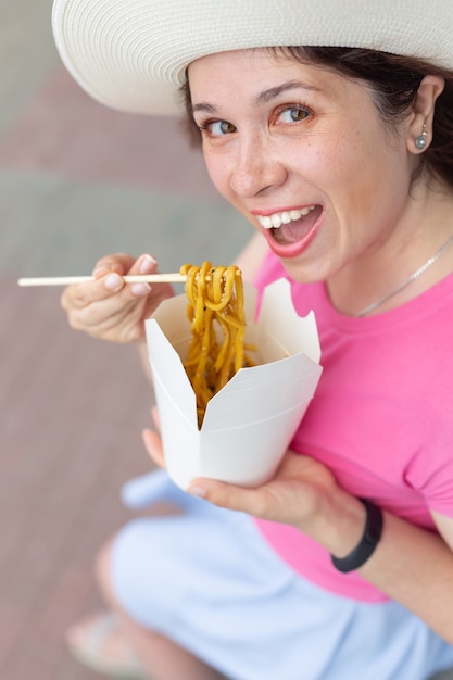 Nahaufnahme der jungen Frau in einem Hut isst chinesische Nudeln, während im Park an einem warmen Sommertag spazieren geht. Food-Konzept zum Mitnehmen.