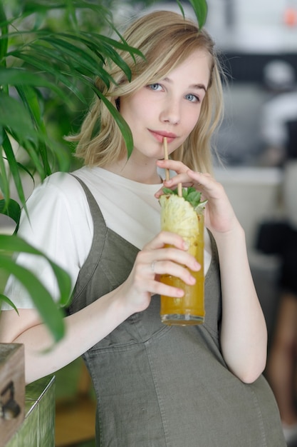 Nahaufnahme der jungen, fröhlichen Frau im Kleid, die glücklich in die Kamera schaut, mit großen grünen Blättern im Hintergrund im gemütlichen grünen Café. Mädchen mit Brille. einen Drink in den Händen halten