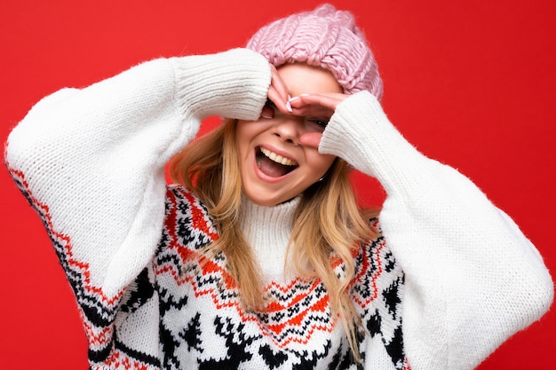 Nahaufnahme der jungen positiven glücklichen schönen blonden Frau mit aufrichtigen Gefühlen, die rosa Strickmütze und Winterpullover einzeln auf rotem Hintergrund trägt und Brille mit den Händen zeigt.