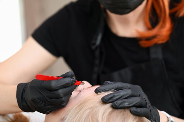 Nahaufnahme der Kosmetikerin, die Handschuhe trägt und Augenbrauen mit einer Pinzette zupft. Salonarbeiterin bereitet Frauenbrauen für dauerhaftes Make-up vor. Mua und Schönheitsbehandlungskonzept