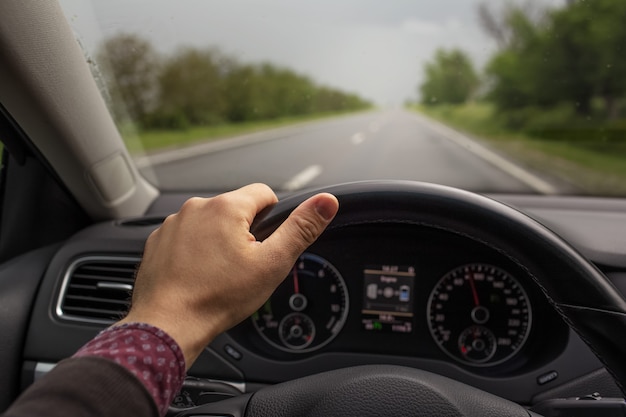 Nahaufnahme der männlichen Hand, die das Lenkrad hält und das Auto fährt. Hintergrund der verschwommenen Straße am regnerischen Tag. Reisekonzept.