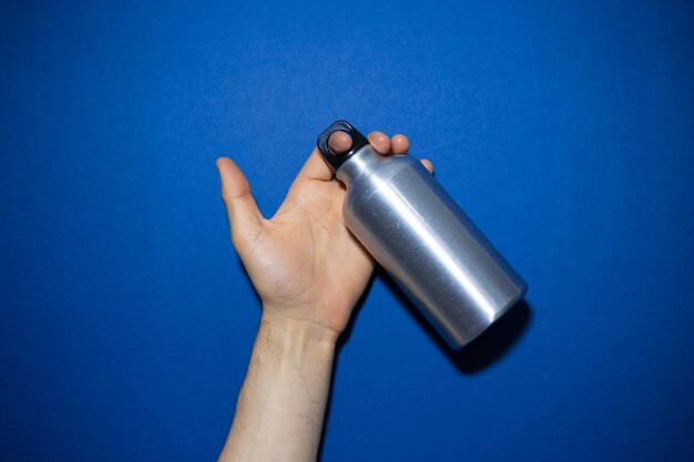 Nahaufnahme der männlichen Hand, die wiederverwendbare Aluminium-Thermowasserflasche hält, die auf Phantomblau der Farbe isoliert wird.